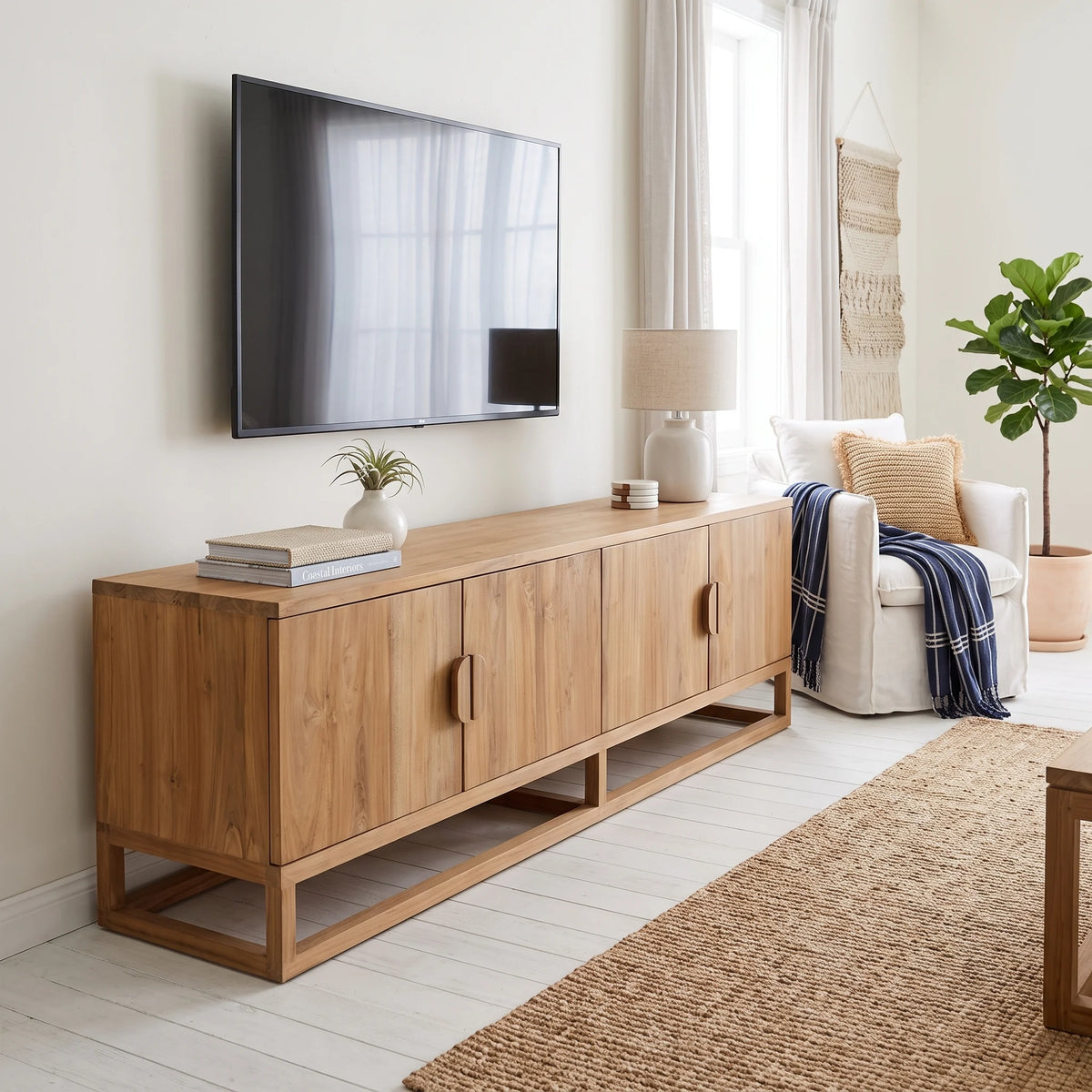 A mid-century modern wooden TV console in a warm oak finish, styled in a bright living room with a wall-mounted TV, a white armchair, and a potted plant.