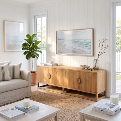 A light natural wood sideboard cabinet in a bright, modern coastal-themed living room. The cabinet features four doors with round pull handles, an open lower shelf, and is styled with books, a vase, and a small plant.