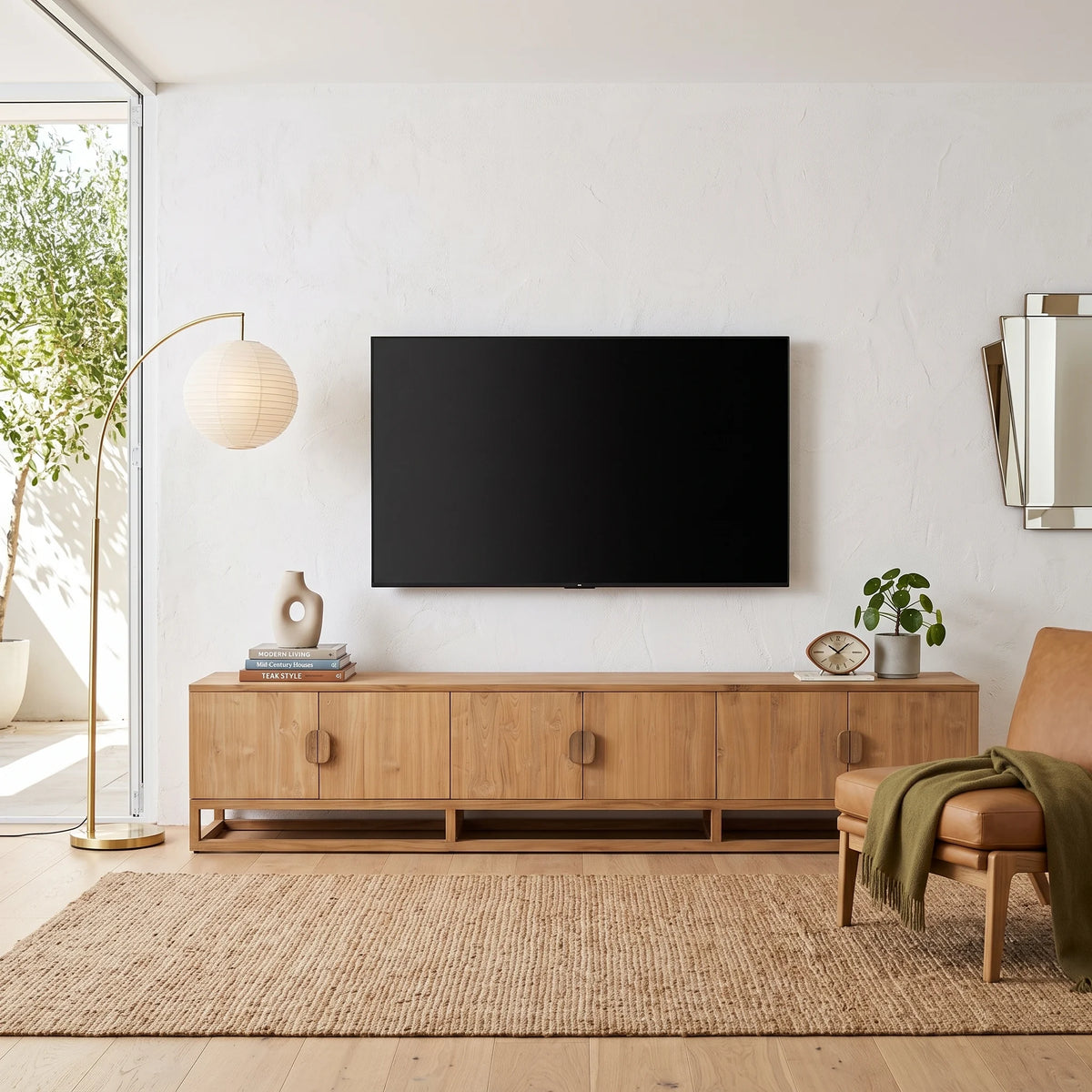 A modern living room scene featuring a light oak wood TV stand with round pull knobs, a black flat-screen TV mounted on a white wall above it, a floor lamp, a chair with a green throw, and a woven rug.