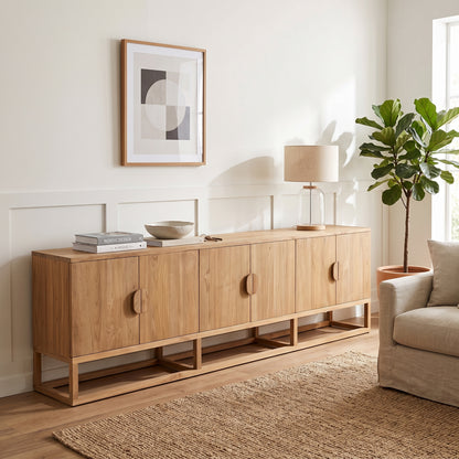 A light teak wood sideboard or credenza with five cabinet doors and open lower shelves, styled in a bright living room with a lamp, framed wall art, a plant, and a woven rug.