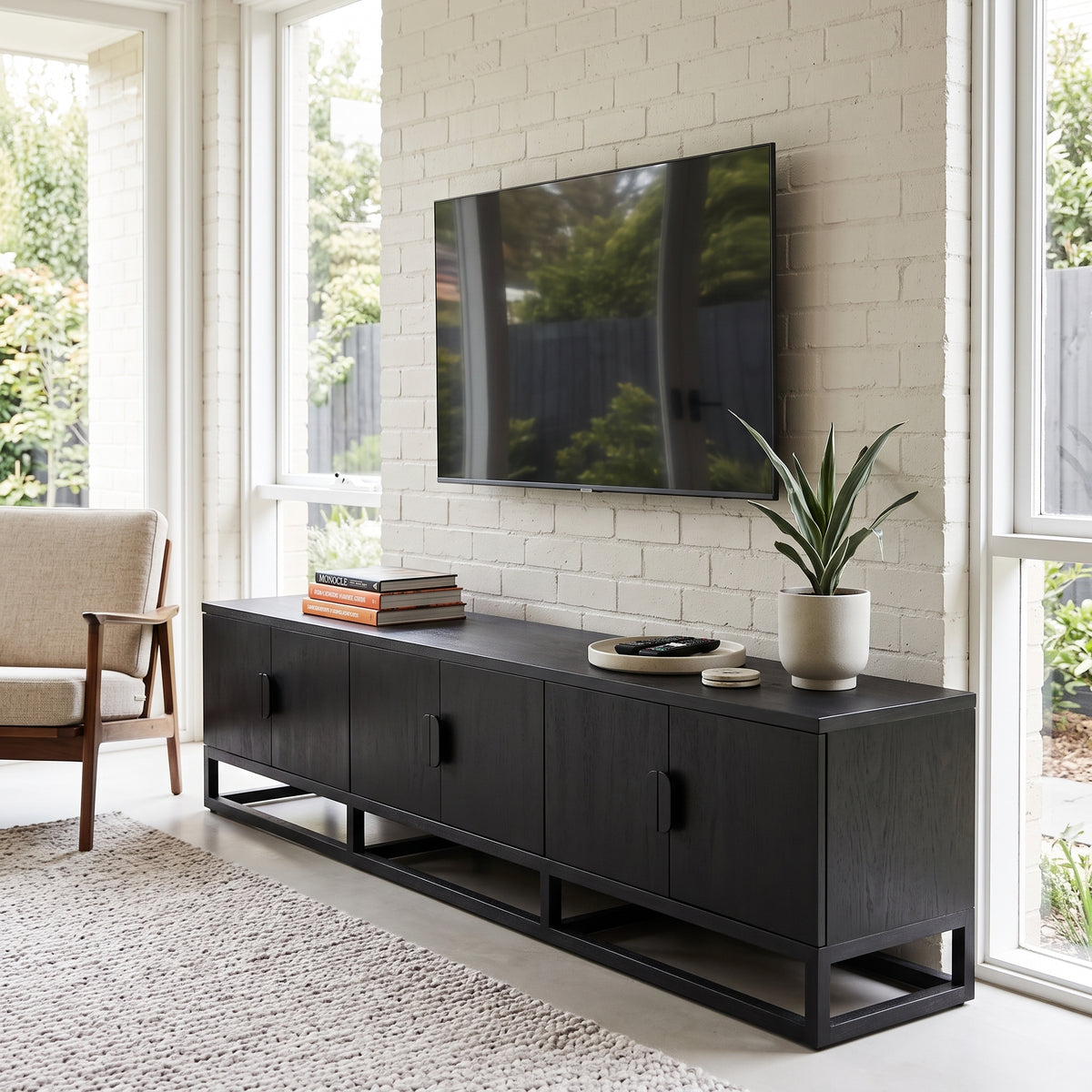 A modern black wooden media console with open side cabinets and a metal frame base, styled in a bright living room with a TV on top, books, a plant, and a chair nearby.