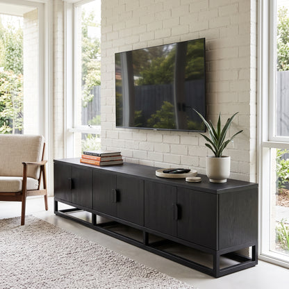 A modern black wooden media console with open side cabinets and a metal frame base, styled in a bright living room with a TV on top, books, a plant, and a chair nearby.