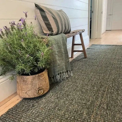 Green textured rug with a wooden stool, plant, and striped pillow against a white wall.