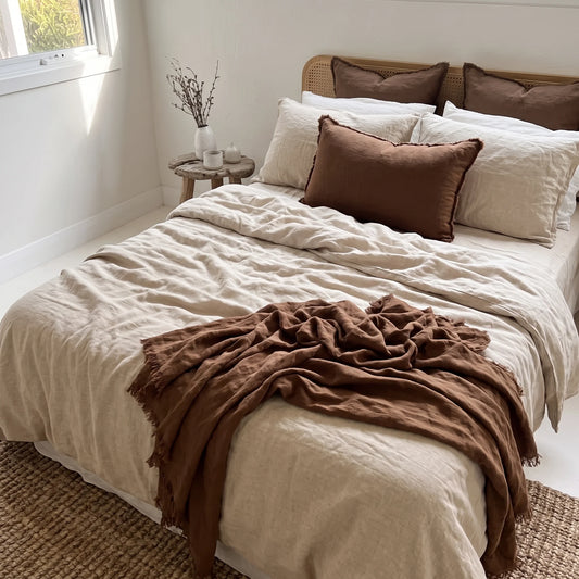 A neatly made bed in a bright bedroom setting, styled with cream-colored bedding, a brown throw blanket draped across the foot of the bed, and several brown and cream pillows. A small wooden side table with a vase of branches sits beside the window.