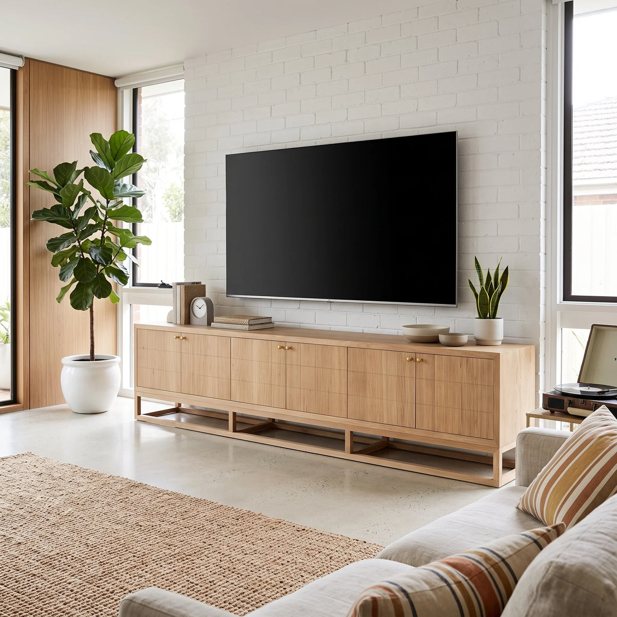 A modern living room scene featuring a light natural wood sideboard console with a large black flat-screen TV mounted above it against a white brick wall. The room includes a large potted fiddle-leaf fig, a small potted plant, decorative bowls, a clock, and a woven rug in the foreground.