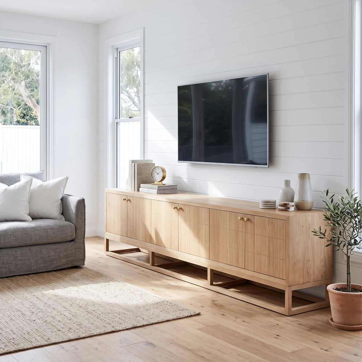 A light natural wood sideboard console cabinet with open shelving and small round knobs, styled in a bright modern living room with a TV mounted on the wall above it.