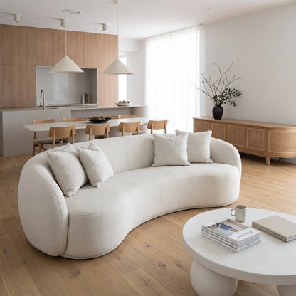 A modern curved upholstered sofa in a bright, minimalist living room with light wood flooring, a matching coffee table, and a kitchen in the background.