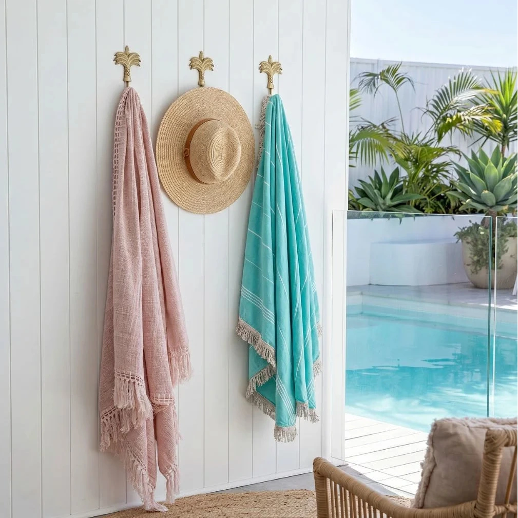 Pink towel, straw hat, and turquoise towel hanging on a white wall with a pool and plants in the background.