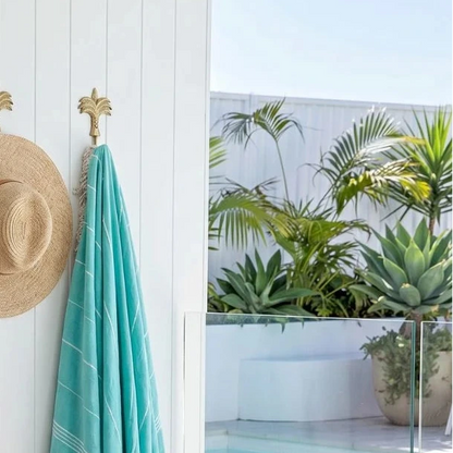 Pink towel, straw hat, and turquoise towel hanging on a white wall with a pool and plants in the background.