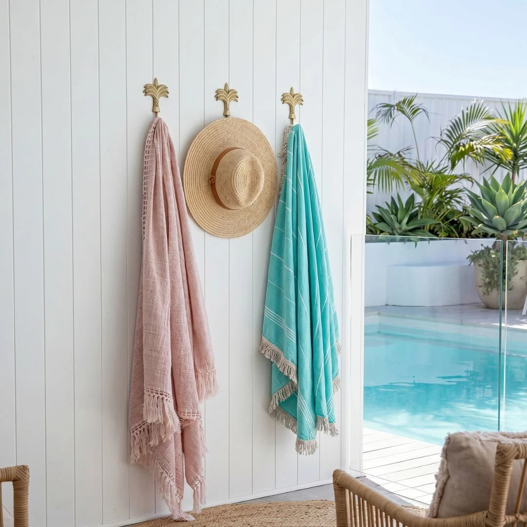 Pink towel, straw hat, and turquoise towel hanging on a white wall with a pool and plants in the background.