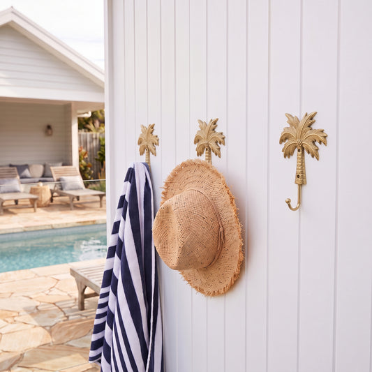 Three gold palm tree wall hooks mounted on a white vertical siding wall, with a navy and white striped towel and a straw hat hanging from two of the hooks, with a pool and patio visible in the background.