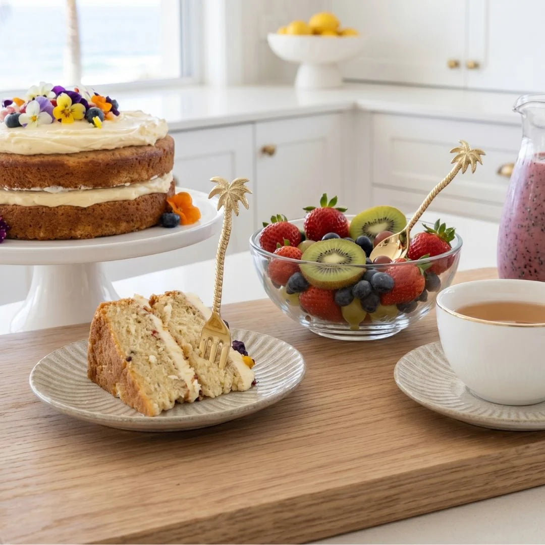 Naked cake with a slice on a plate, fruit bowl, and cup of coffee on a wooden table.