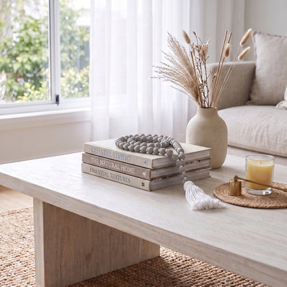 A styled interior scene showing a light wood coffee table in a bright living room. On the table are stacked books, a gray beaded necklace, a small white object, and a candle on a woven coaster.