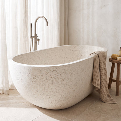 A freestanding oval bathtub in a warm beige speckled stone or concrete finish, shown in a softly lit bathroom with a chrome faucet, sheer curtains, a small wooden stool, and a draped towel.