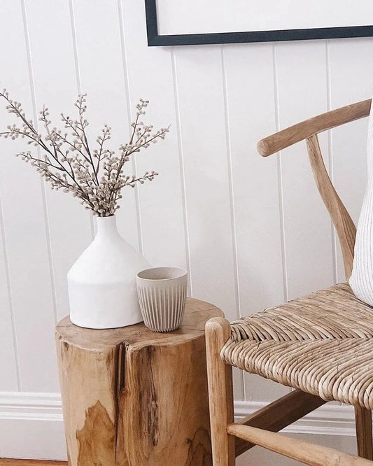Wishbone chair with a cushion next to a teak stump side table with a vase and small cup against a white paneled wall.