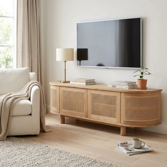 A light natural wood TV stand with woven cane panels, styled in a bright living room with a wall-mounted TV, a white armchair, and a lamp.