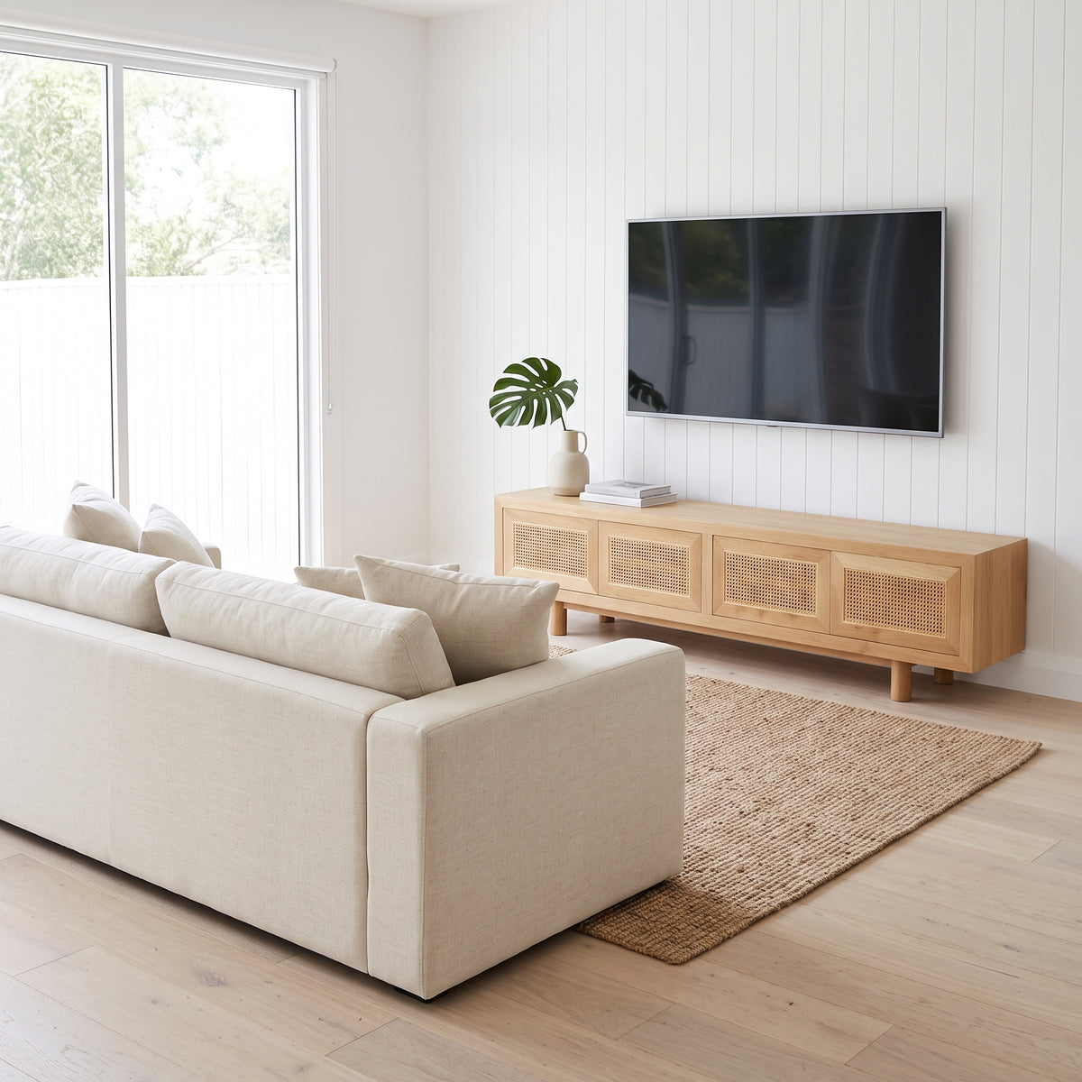 A modern living room scene featuring a light beige fabric sofa, a natural wood TV stand with woven cane doors, a flat-screen TV, a woven jute rug, and a white shiplap wall.
