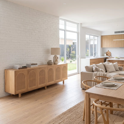 A light natural wood sideboard with arched cane panel doors, styled in a bright, modern living room with a white brick wall, wooden floors, and a dining table nearby.
