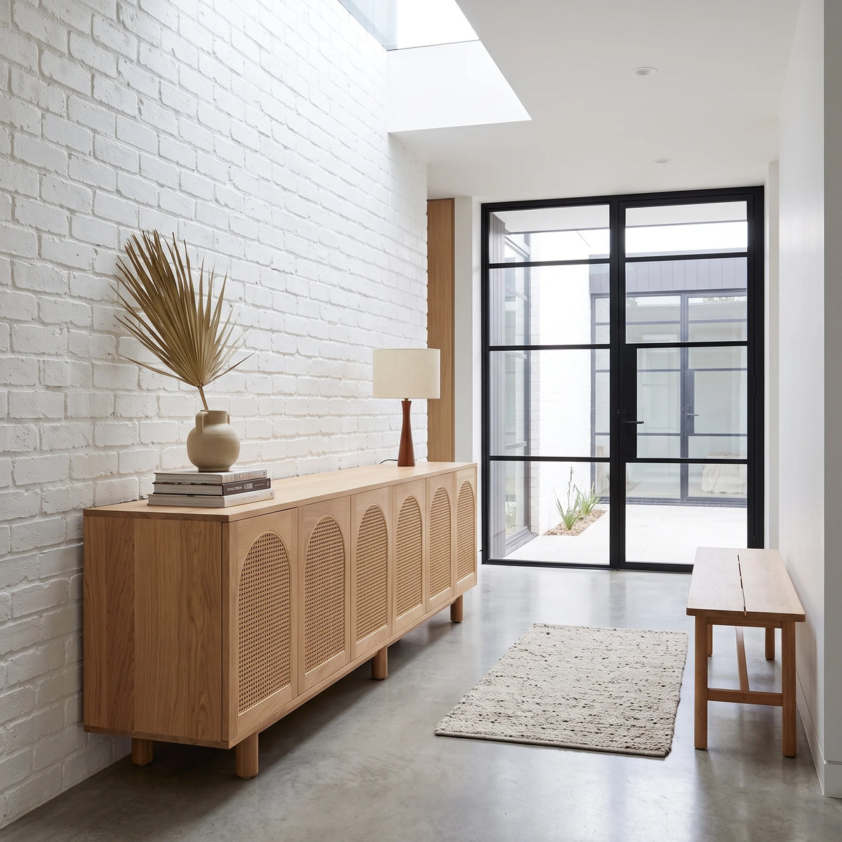 A light natural wood credenza with arched slatted doors stands against a white brick wall in a bright, modern entryway. The credenza is styled with a stack of books, a ceramic vase with dried palm leaves, and a table lamp.