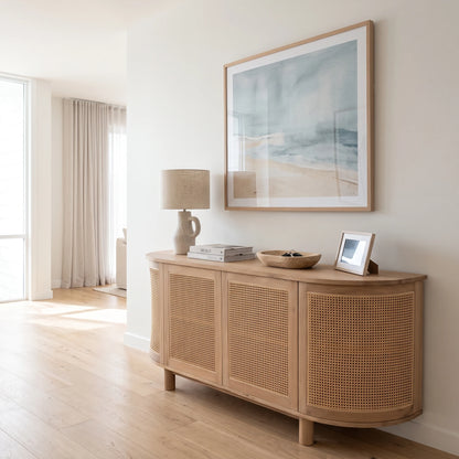A light natural wood sideboard cabinet with woven cane panels, styled in a bright, modern room with a lamp, books, and a framed print on top, beneath a large framed beach art print.