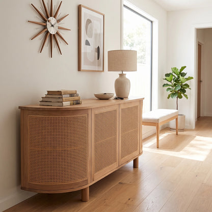 A light natural wood sideboard cabinet with woven cane panel doors, styled in a bright, modern room with a lamp, books, and a bench.