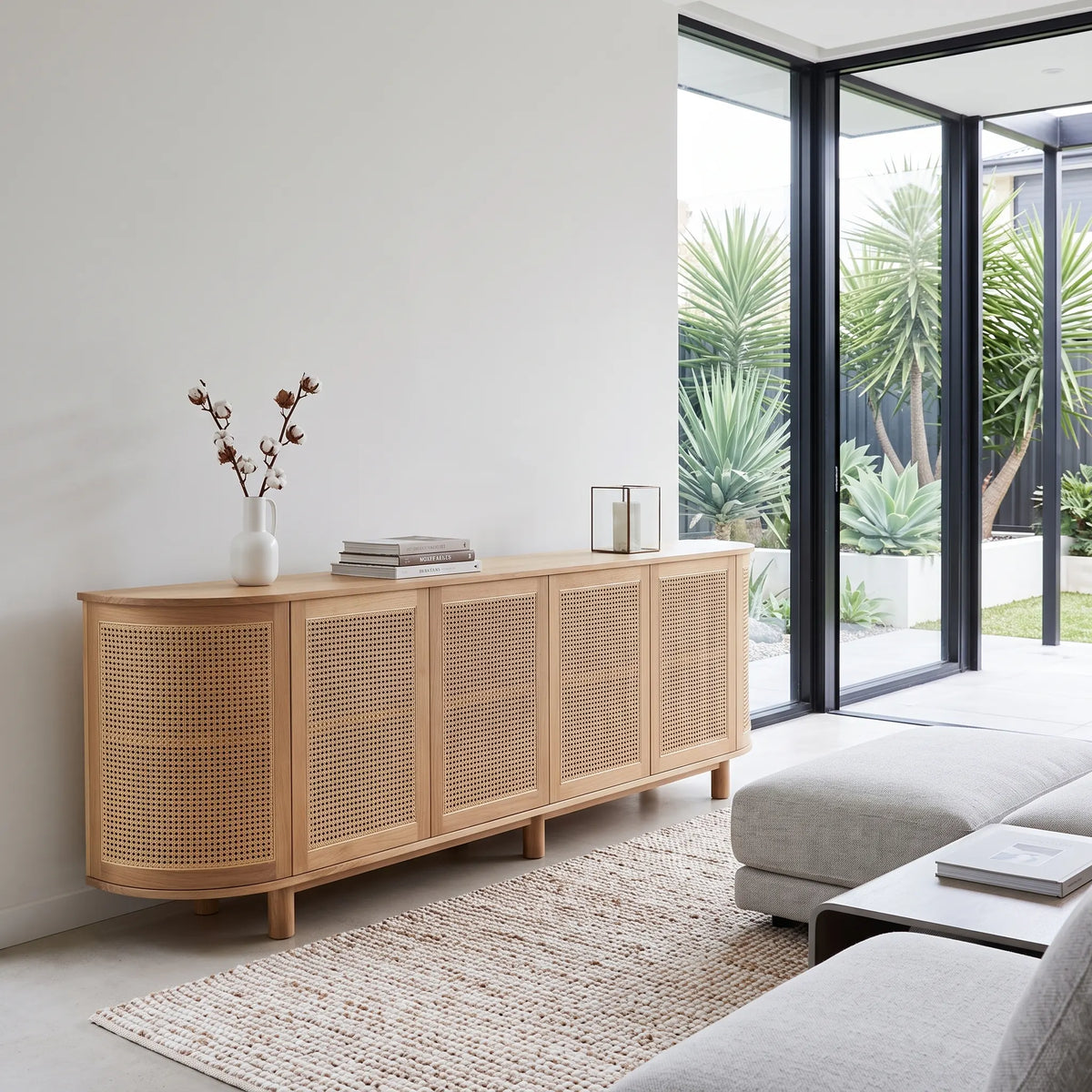 A light natural wood sideboard with woven cane panels, styled in a modern minimalist living room next to a glass patio door.