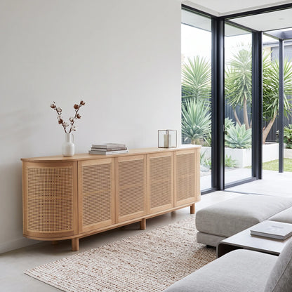 A light natural wood sideboard with woven cane panels, styled in a modern minimalist living room next to a glass patio door.