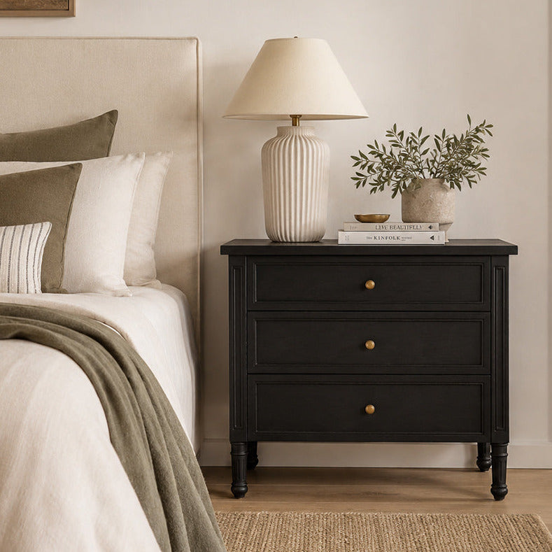A black three-drawer nightstand in a styled bedroom setting, styled with a ribbed ceramic table lamp, a small potted plant, and books on top.
