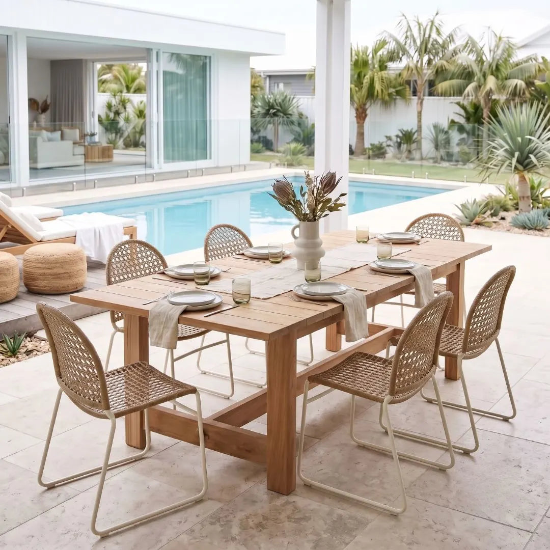 An outdoor dining set on a patio beside a swimming pool, featuring a rectangular wooden table with a woven rattan-style top and six matching chairs with metal frames, set with plates and a vase of flowers.