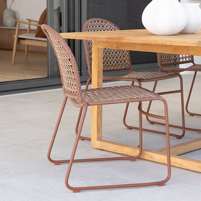A modern dining set featuring a light wood table and woven rattan chairs with a warm rust-colored metal frame, set on a light concrete patio.