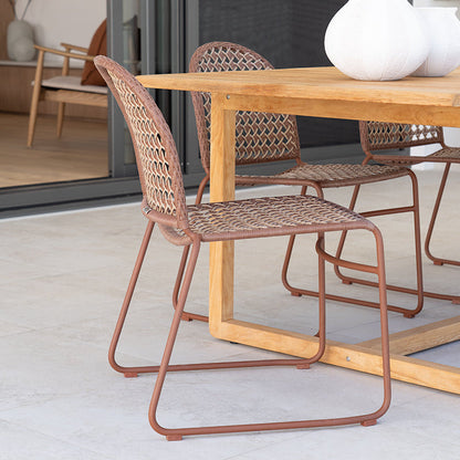 A modern dining set featuring a light wood table and woven rattan chairs with a warm rust-colored metal frame, set on a light concrete patio.