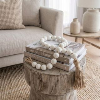 White wooden bead garland with a tassel draped over a stack of books on a rustic wood side table in a living room setting.