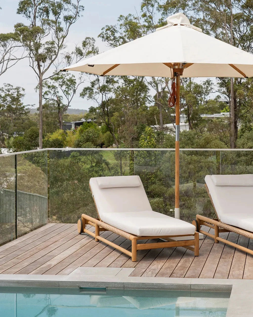 Two lounge chairs with an umbrella on a wooden deck by a pool, surrounded by trees.