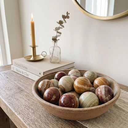Decorative bowl with marbled onxy stones on a wooden surface, candle, books, and plant in the background.
