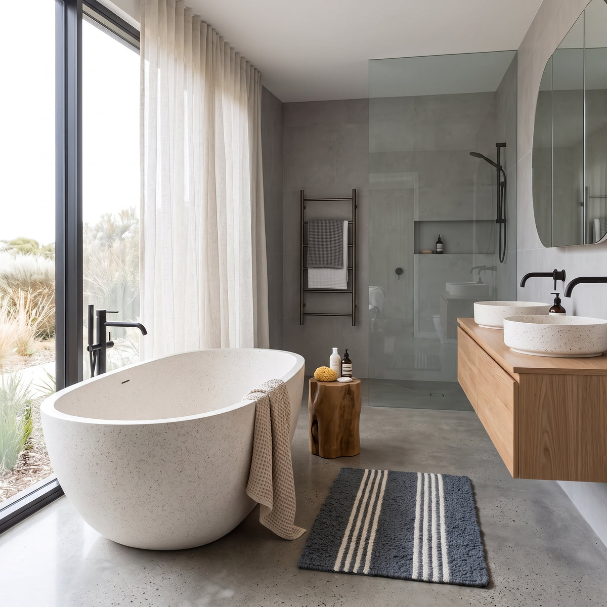 A modern freestanding oval bathtub in a light speckled stone finish, styled in a contemporary minimalist bathroom with a wood floating vanity, double white vessel sinks, a glass shower enclosure, and a dark blue striped bath mat.