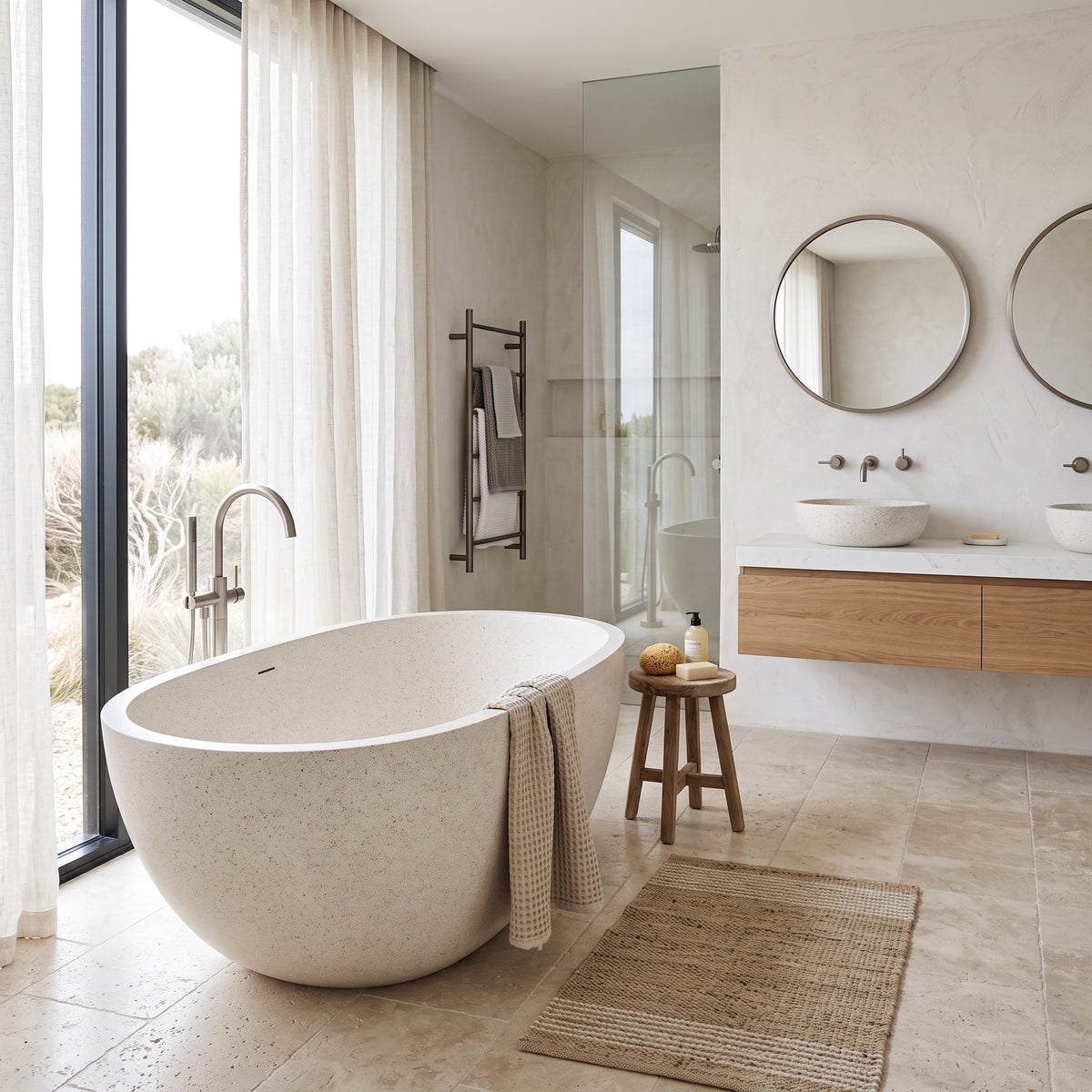 A modern freestanding oval bathtub in a light speckled stone finish, styled in a contemporary beige-toned bathroom with a wooden vanity, round mirrors, and a large window looking out onto a desert landscape.