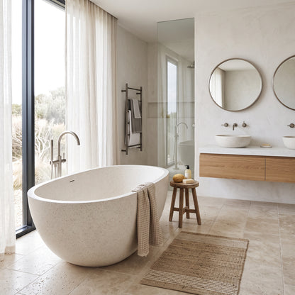 A modern freestanding oval bathtub in a light speckled stone finish, styled in a contemporary beige-toned bathroom with a wooden vanity, round mirrors, and a large window looking out onto a desert landscape.