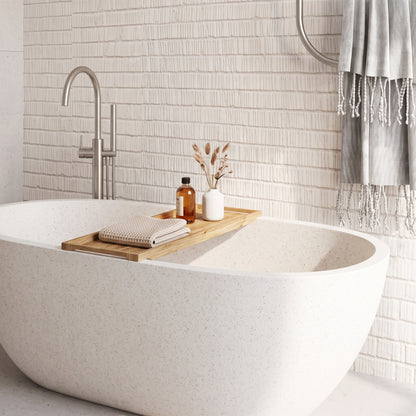 A freestanding oval bathtub in a bright bathroom with a textured white tile wall, a silver faucet, and a wooden bath tray holding a folded towel, amber bottle, and small vase with dried stems.