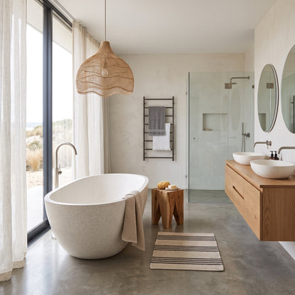 A modern bathroom interior featuring a freestanding oval white bathtub, a wicker pendant light, a wooden floating vanity with two white vessel sinks, a glass shower enclosure, and a concrete floor.