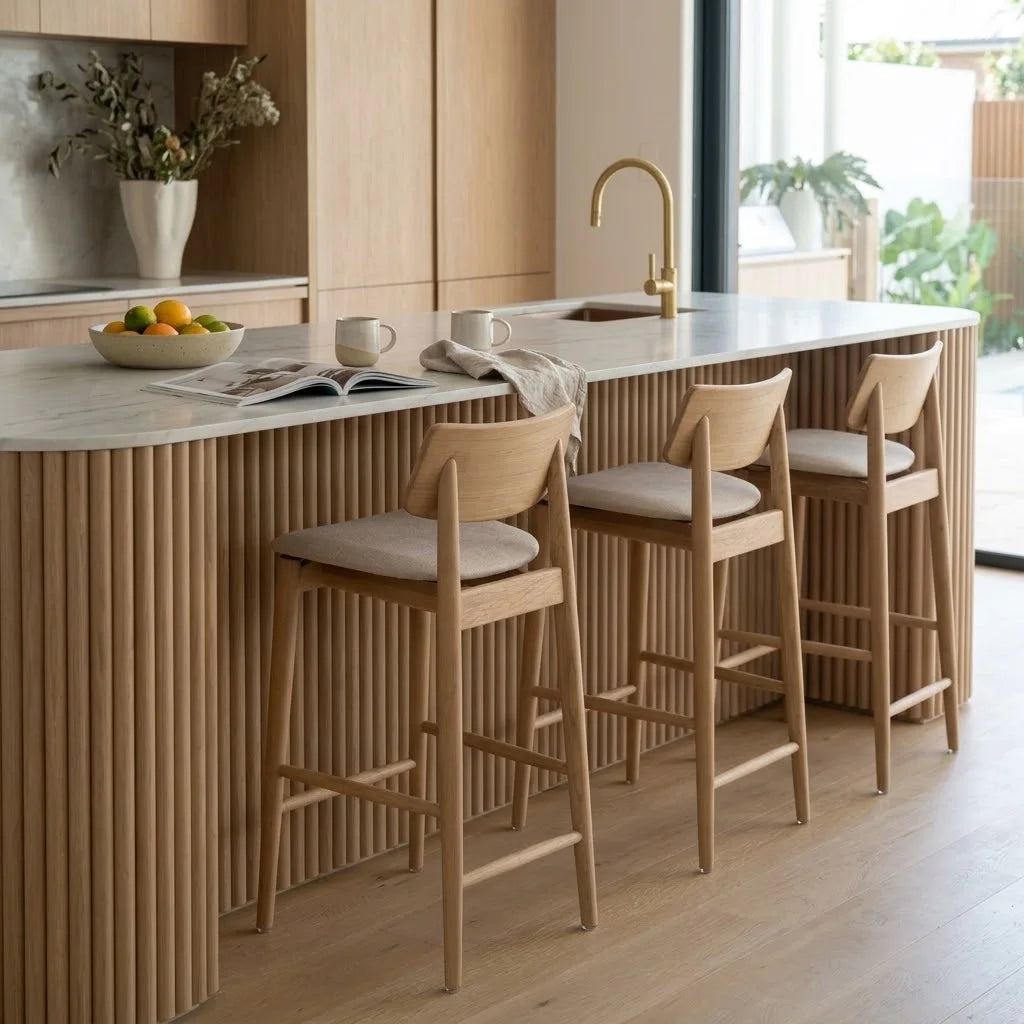 Three wooden bar stools with light beige upholstered seats are arranged along a modern kitchen island with a fluted wood base and white countertop. The kitchen features light wood cabinetry, a white marble-like countertop, a gold faucet, and a window with plants visible outside.