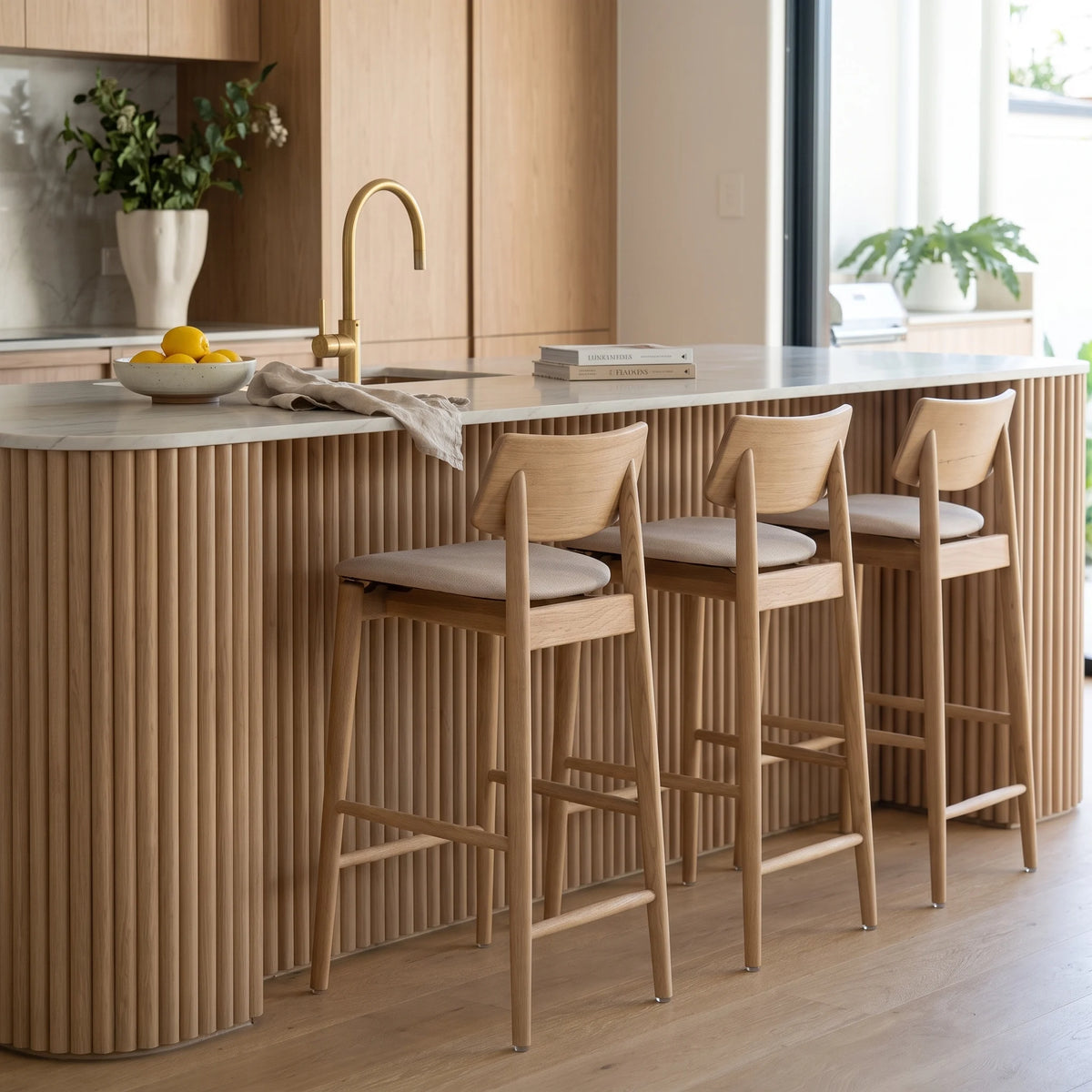 Three wooden bar stools with light gray upholstered seats are arranged along a modern kitchen island with a fluted wood facade, marble countertop, and gold faucet in a bright interior.
