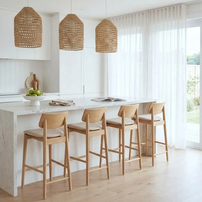 A bright modern kitchen with three woven wicker pendant lights hanging over a white marble island. Four light wood bar stools with beige cushions sit at the counter.
