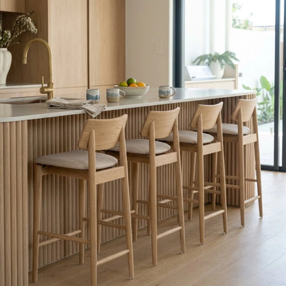 Four light oak wood bar stools with beige upholstered seats arranged along a modern kitchen island with a slatted wood front and a white countertop.