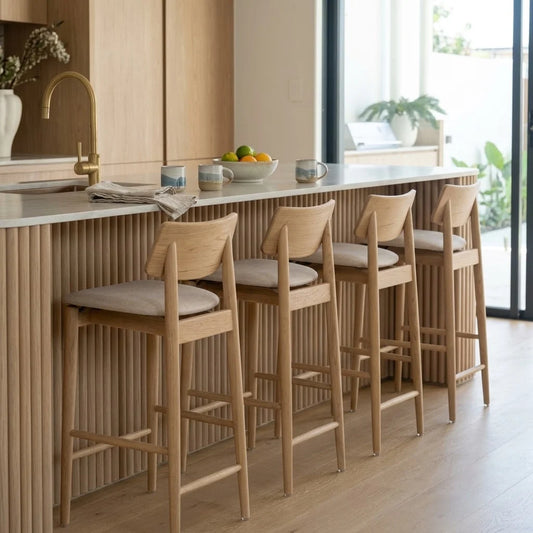 Four light oak wood bar stools with beige upholstered seats arranged along a modern kitchen island with a slatted wood front and a white countertop.