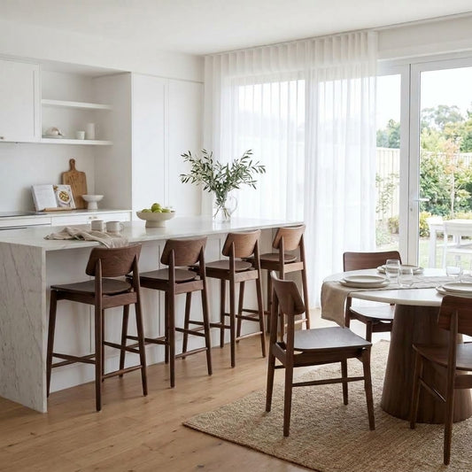 A bright, modern kitchen and dining area with a white marble island, dark wood bar stools, a round dining table, and light wood chairs, set in a sunlit room with sheer white curtains and a view of greenery outside.
