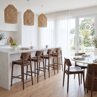 A bright, modern kitchen and dining area with a white marble island, five dark wood bar stools, three woven wicker pendant lights, and a round dining table with wooden chairs near large windows looking out to a garden.