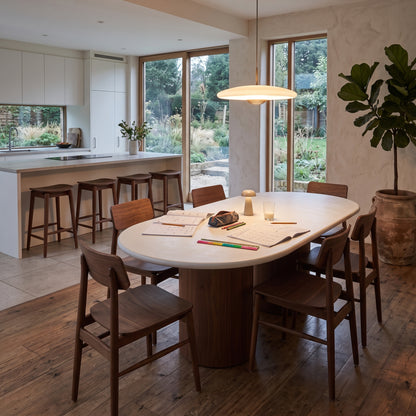 A modern dining room scene featuring a round white dining table with dark wood chairs, set in a bright, contemporary kitchen with large windows and a potted fiddle-leaf fig plant.