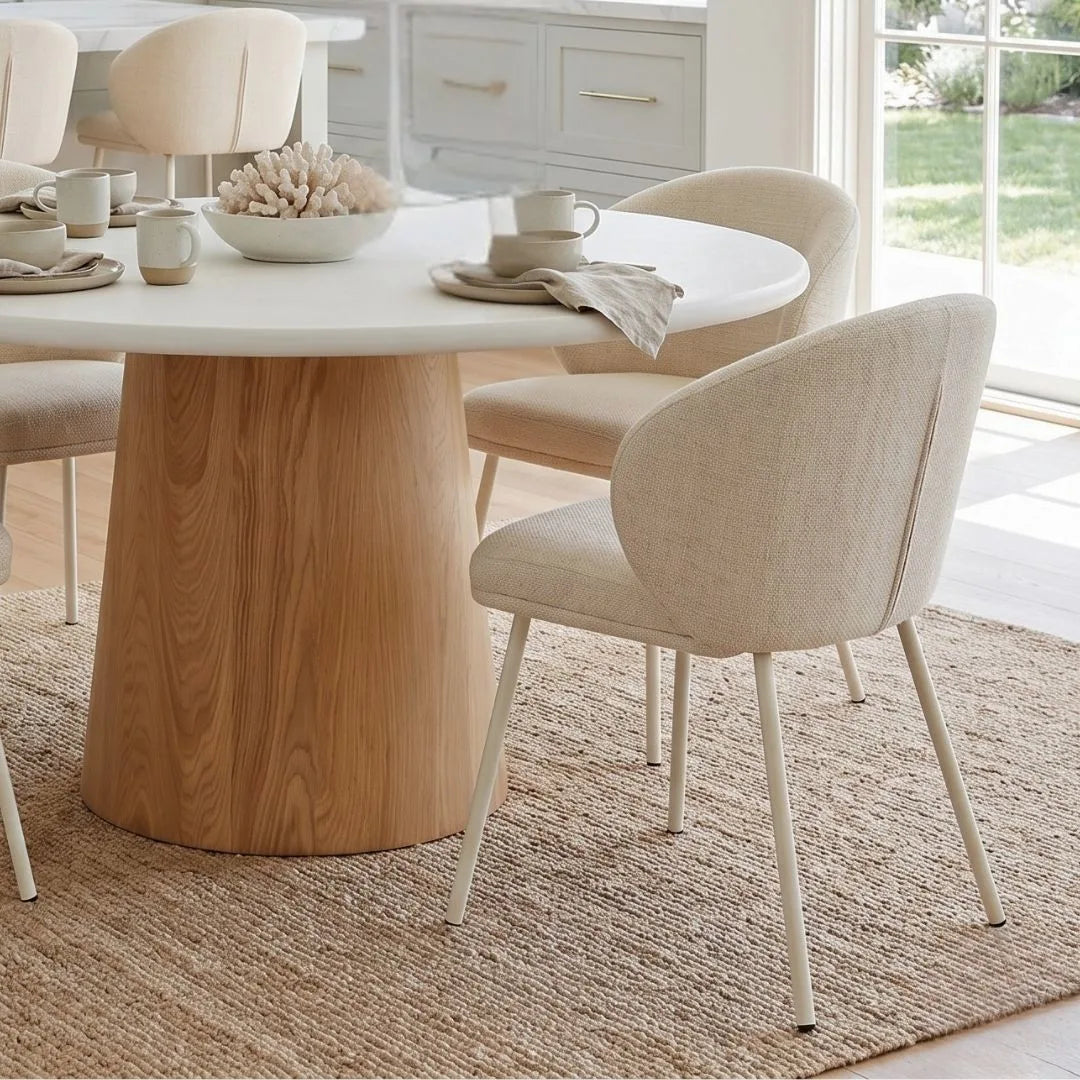 Close up of a the round microcement dining table with natural oak base, linen dining chairs in an modern coastal open plan kitchen dining home by sun republic