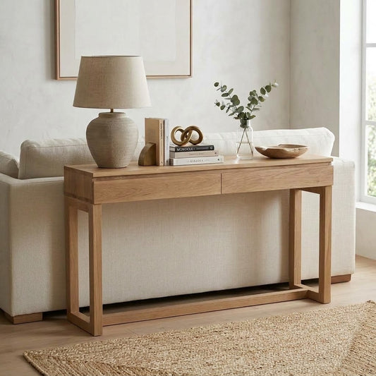 A light natural oak console table styled in a bright living room setting, featuring a ceramic table lamp, books, decorative objects, and a small vase on top.