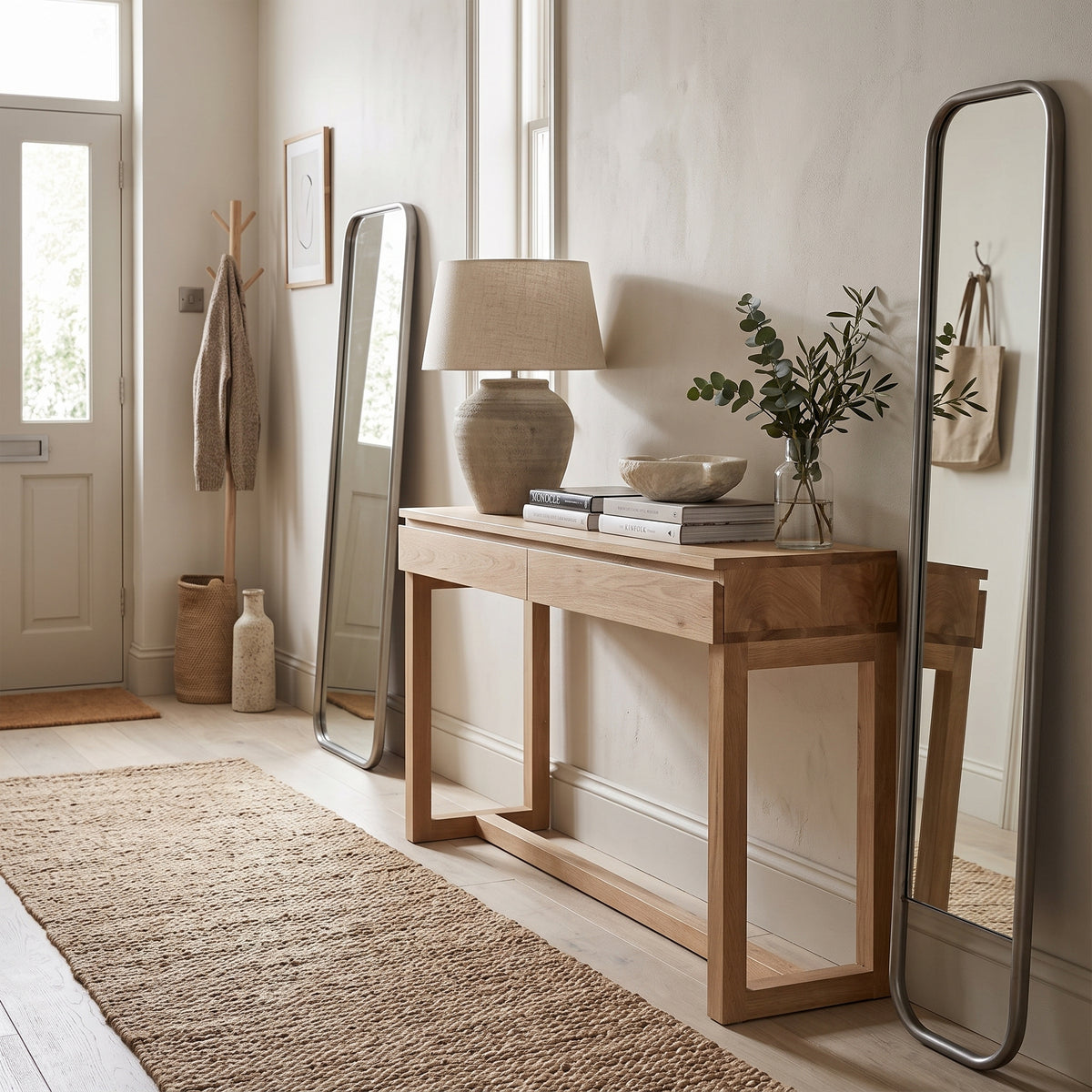 A light natural wood console table styled in a bright, minimalist room with a woven rug, a ceramic table lamp, books, and two tall floor mirrors.